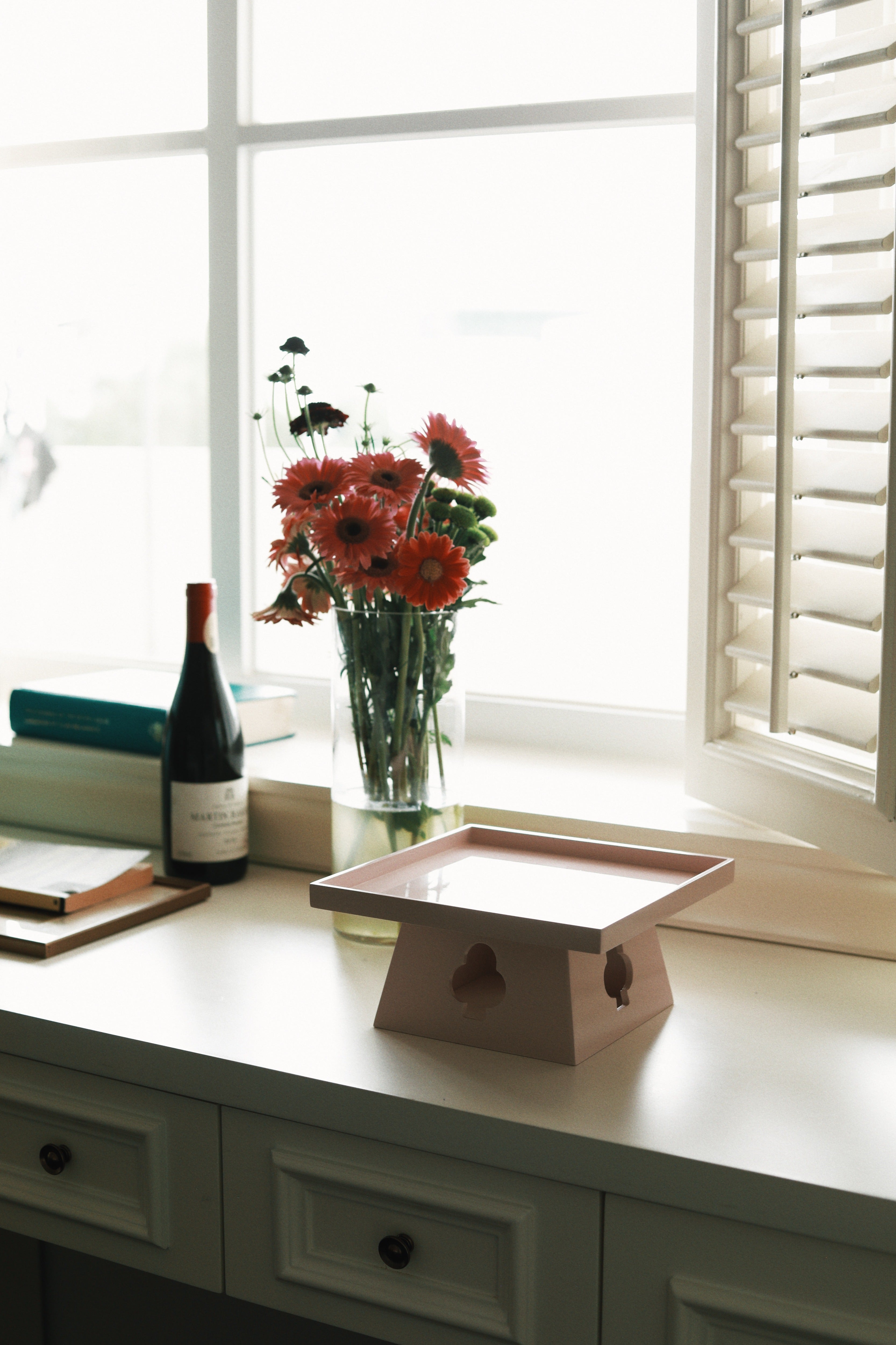 Vinidec elevated tray in dusty pink lacquer with spade cutouts, styled on a white desk by a window with flowers and a wine bottle